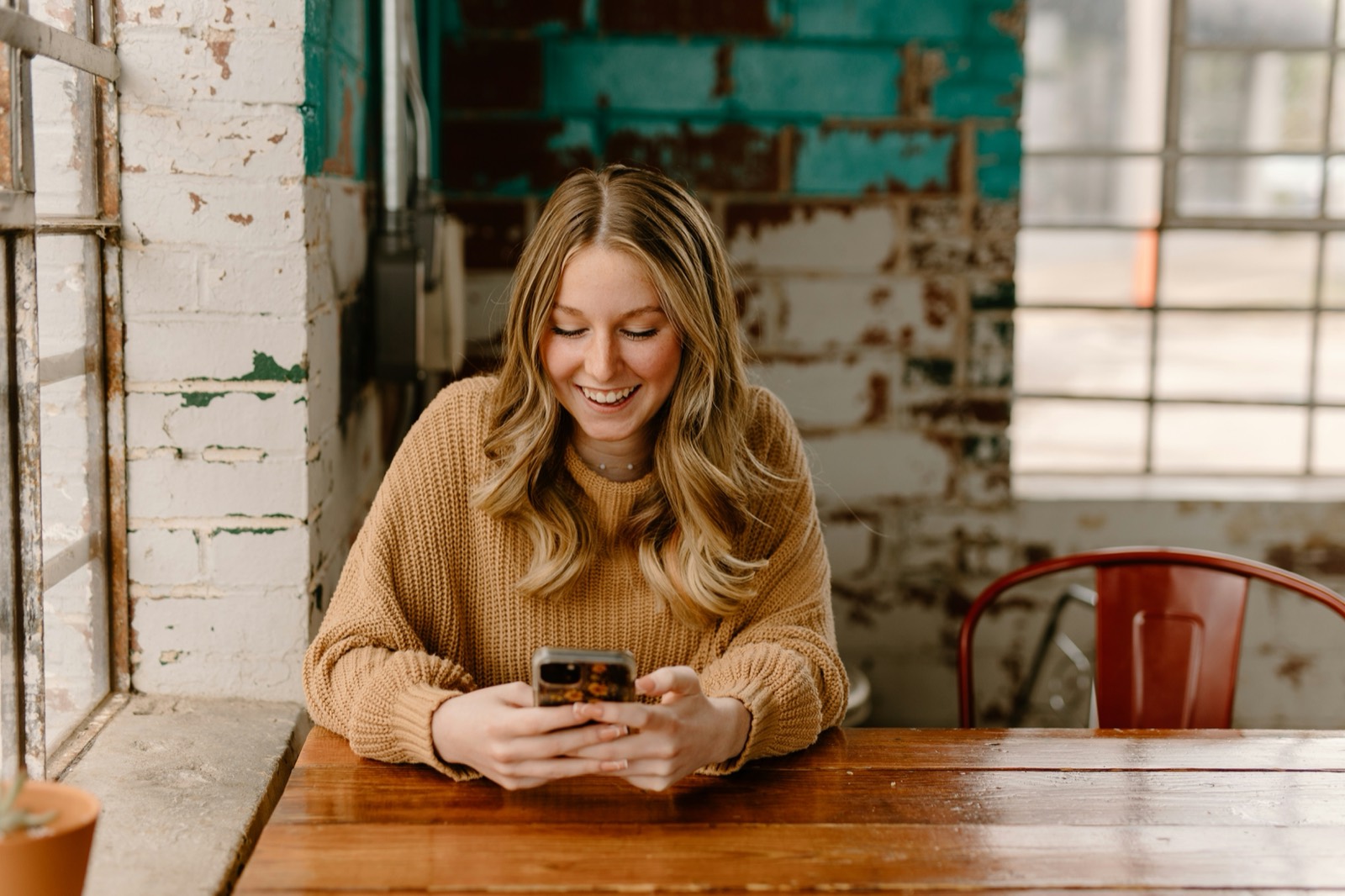 Een jonge vrouw lacht naar haar telefoon aan een tafel in een café